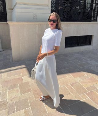 Influencer fesyen asal Swedia Lovisa Barkman berjalan keluar di jalan batu netral mengenakan kacamata hitam cat-eye, kaus putih, rok maxi slip putih, tas dengan pegangan atas putih, dan sandal geser Hermès putih.