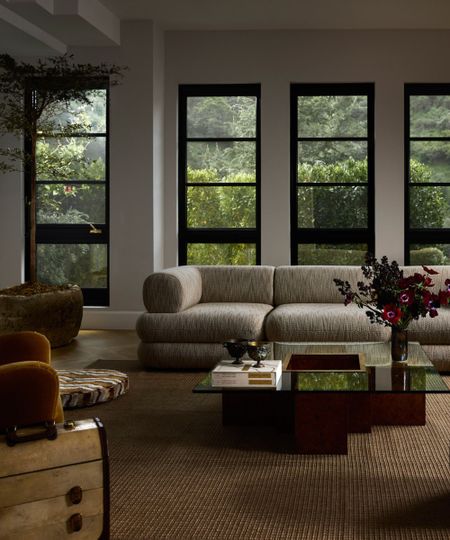 Large modern living room, large cream sofa with yellow armchairs and glass and wood coffee table surrounding, beige carpet and small ottoman, large windows with greenery outside behind