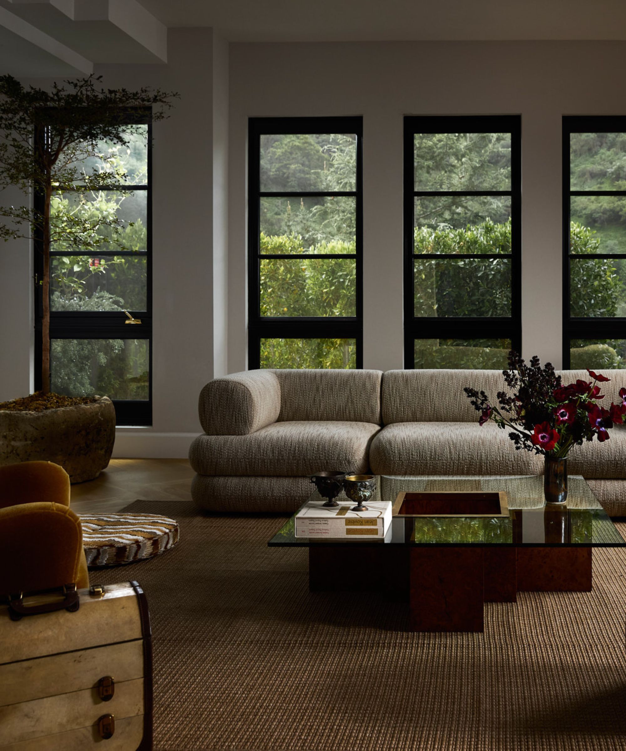 Large modern living room, large cream sofa with yellow armchairs and glass and wood coffee table surrounding, beige carpet and small ottoman, large windows with greenery outside behind