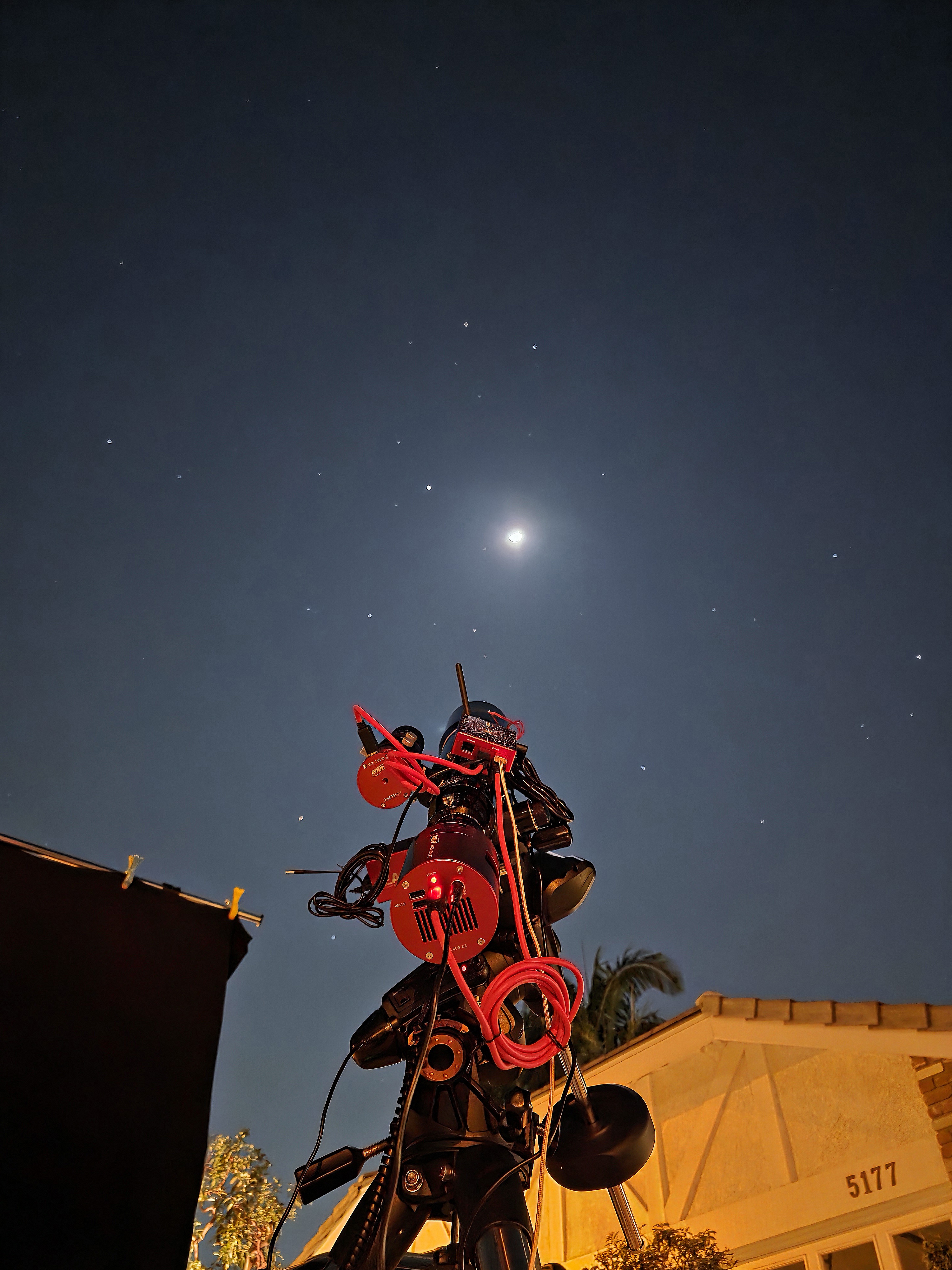 A photo of a telescope pointing at the moon in a starry night sky. The image is taken from behind the telescope and a house is visible at the bottom of the frame.
