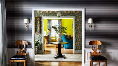 hallway with striped and floral wallpapers and yellow paint, with timber chairs on either side of the door