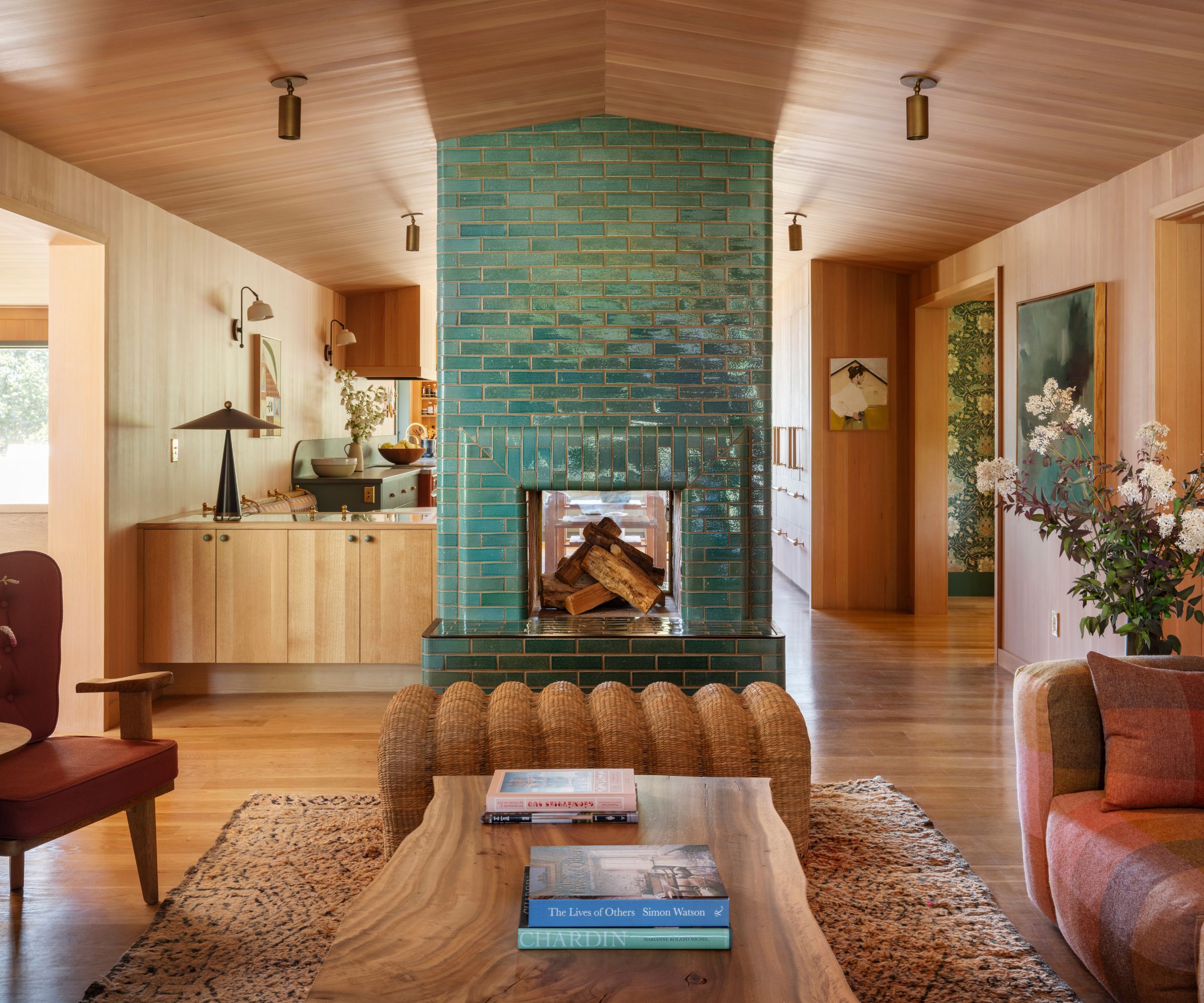 An open-plan living room with a teal brick fireplace, wooden walls, ceiling, and kitchen cabinets.