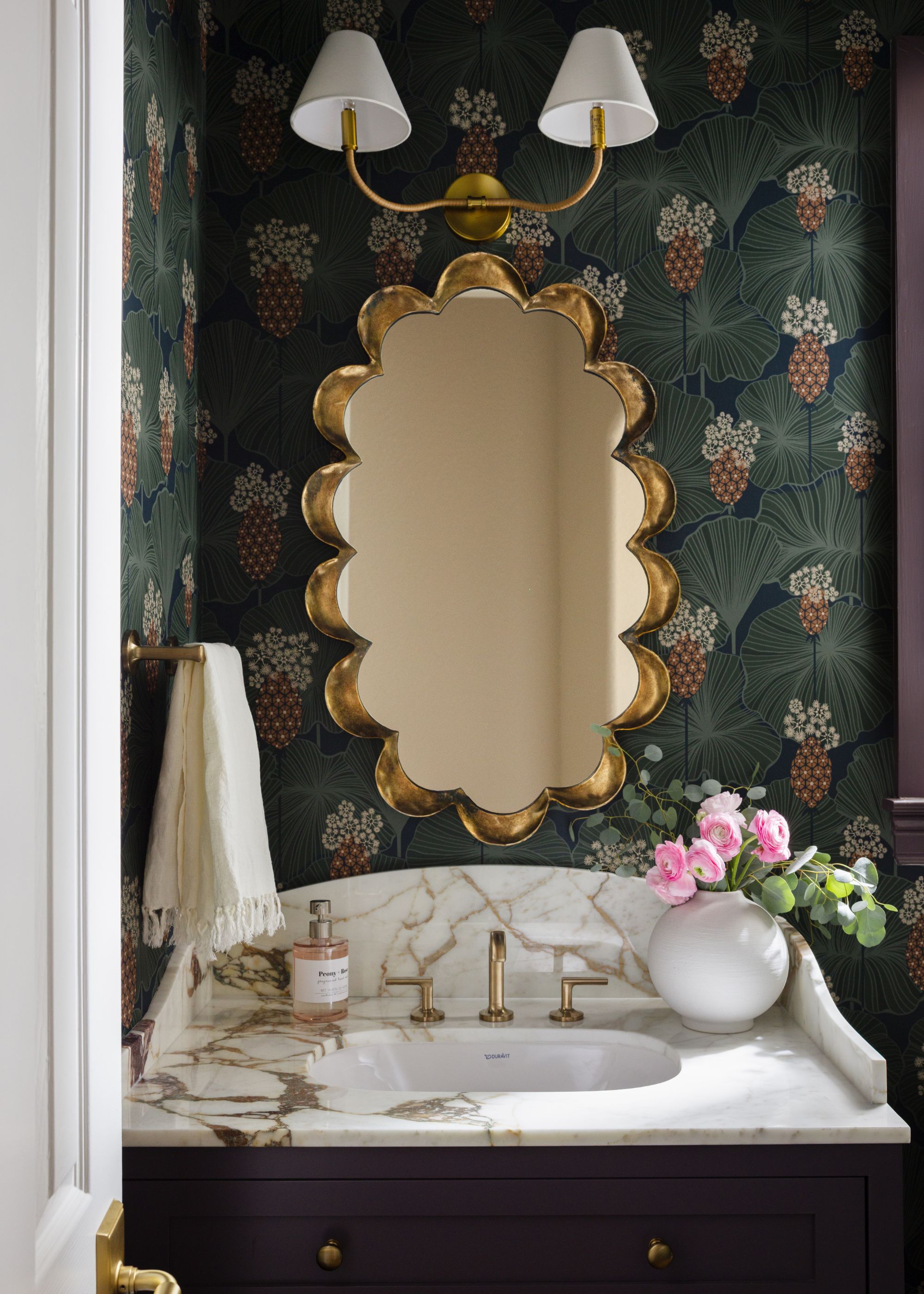 A bold powder room featuring dark floral wallpaper with pineapple motifs. A scalloped gold mirror hangs above a marble-topped vanity with a dark purple base and brass fixtures.