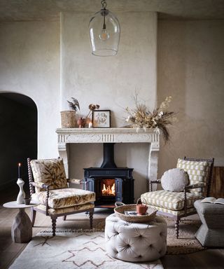 a living room with lit fire in a woodburner, marble mantlepiece and plain marble effect walls, wood floor, two armchairs and dried leaves and grasses floral decorations.