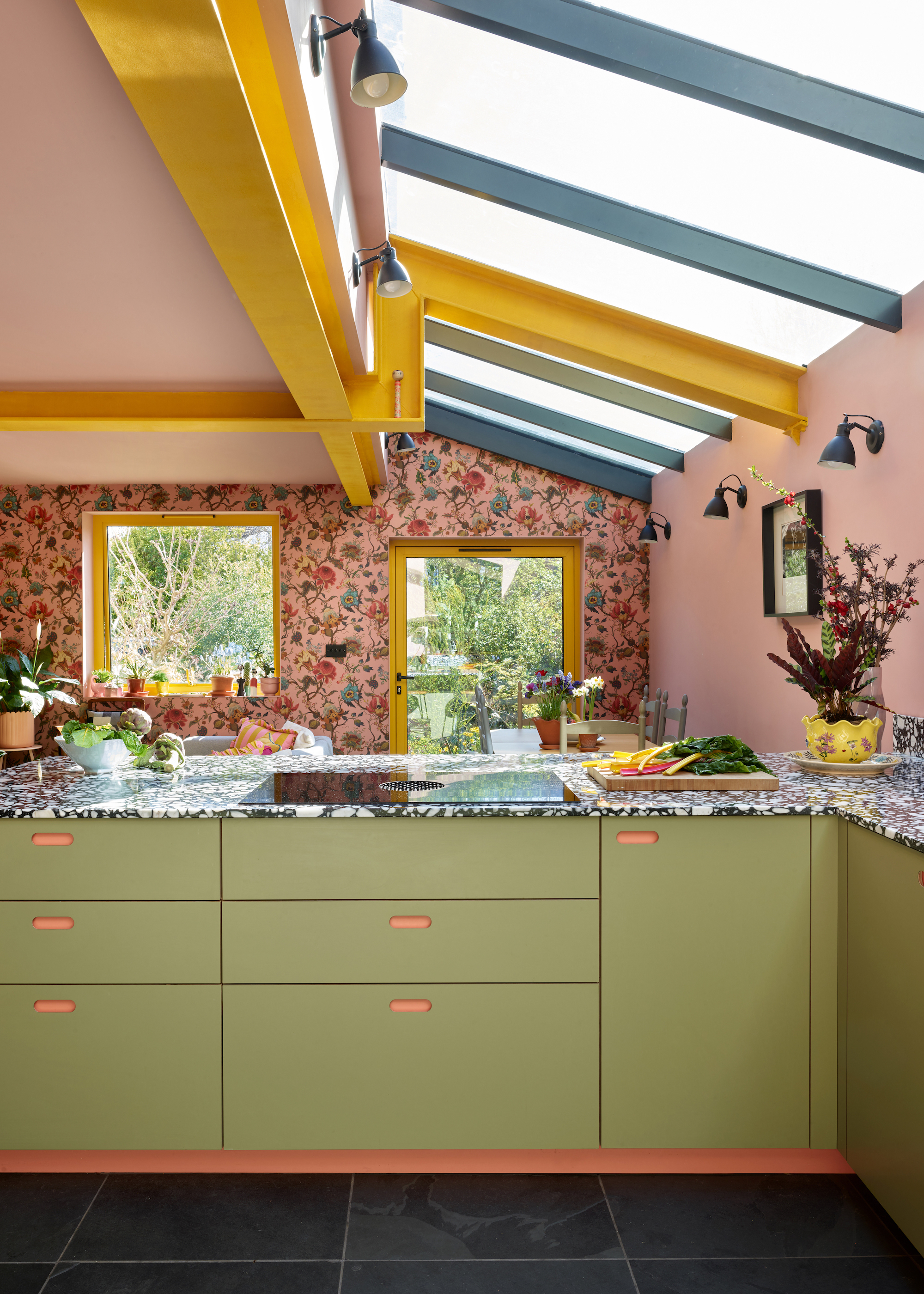 green and coral kitchen with floral wallpaper on the back wall, yellow ceiling beams, skylights, black floor tiles, and yellow trim