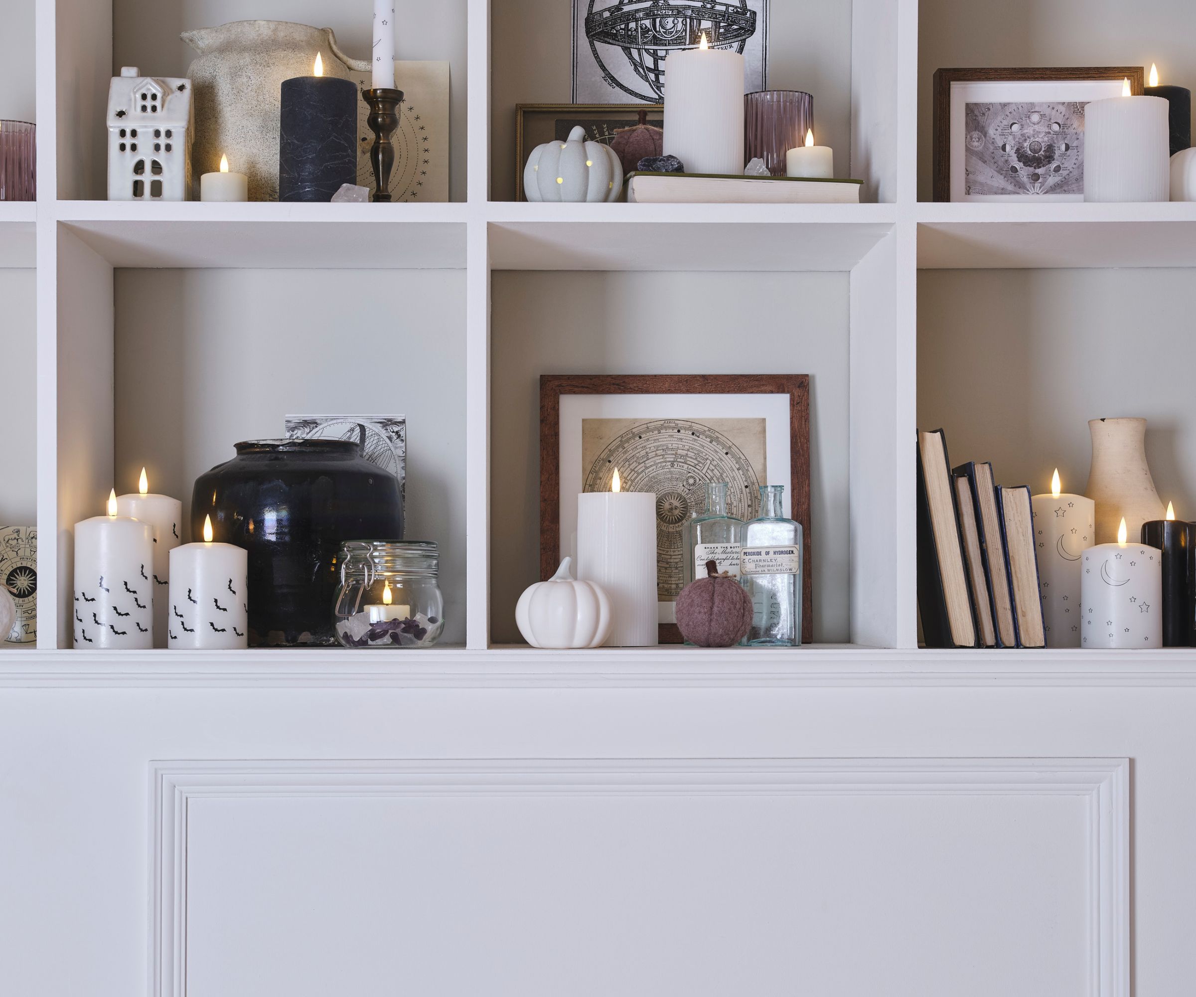 A white built-in shelving unit styled with decorative objects and soft candlelight. Shelves display a mix of framed art, books, ceramic vases, glass jars, and small pumpkins, with glowing candles creating a warm, cosy ambience.