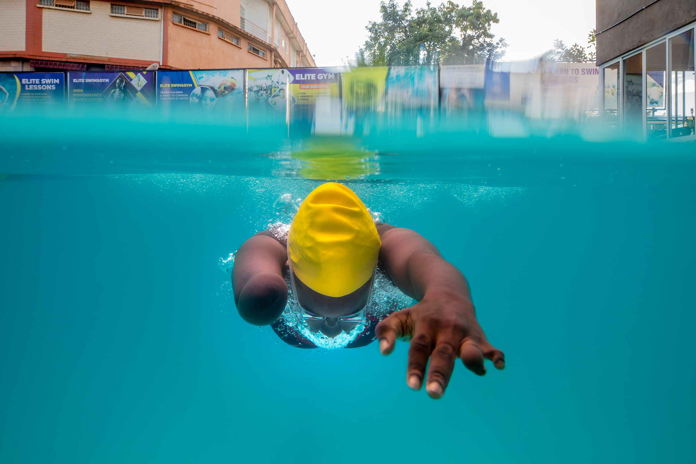 A swimmer in a yellow cap glides through clear blue water, one arm extended forward and bubbles trailing behind
