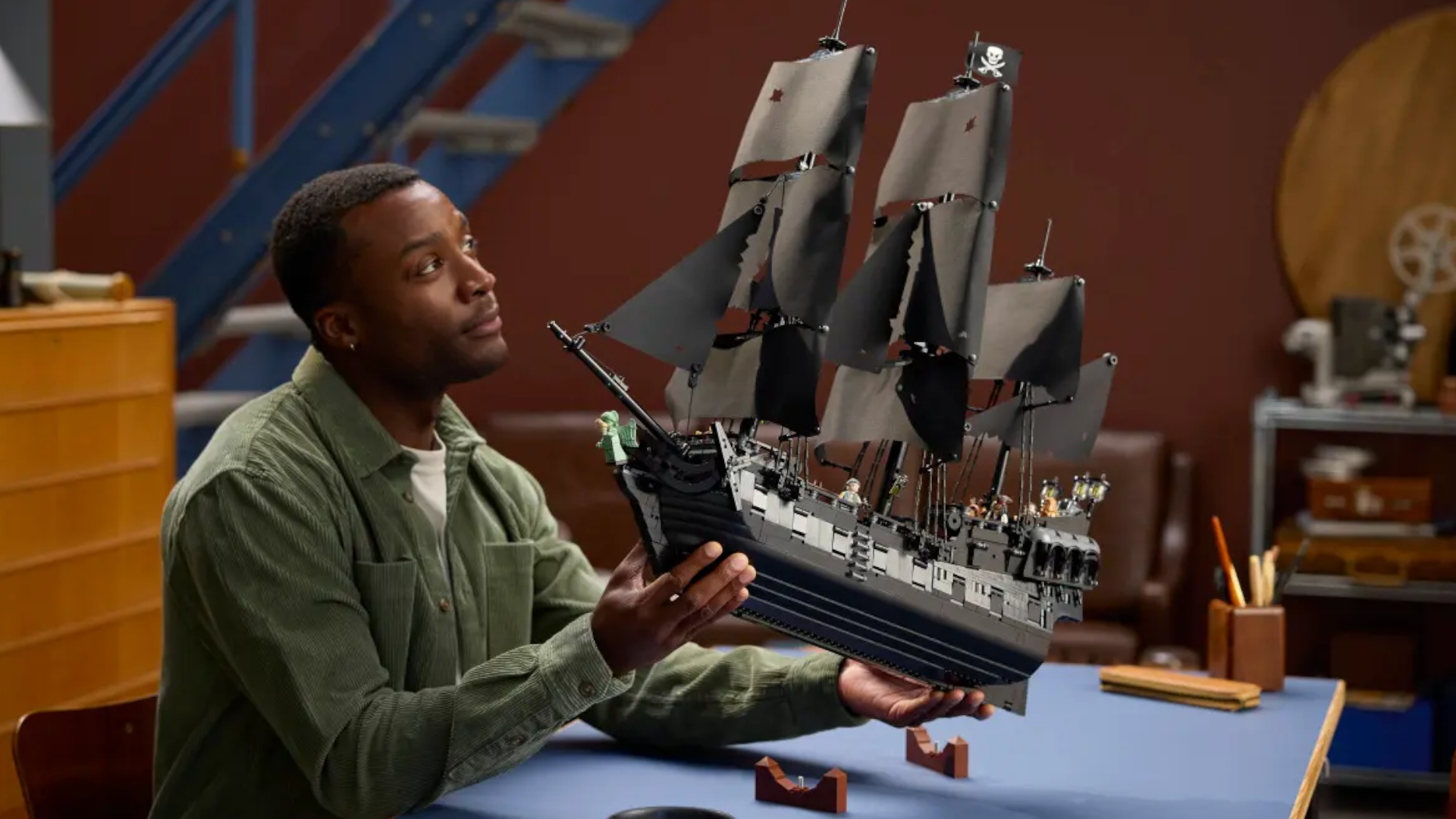 A man holds up the Lego Captain Jack Sparrow&#039;s Pirate Ship model while sitting at a blue table