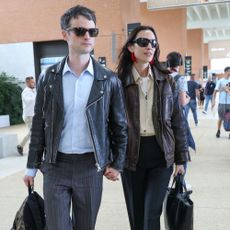 Alexa Chung and Tom Sturridge walk through Venice airport holding hands. Alexa carries a black bag in her hands and wears a brown leather jacket, a yellow shirt, black trousers and black loafers.