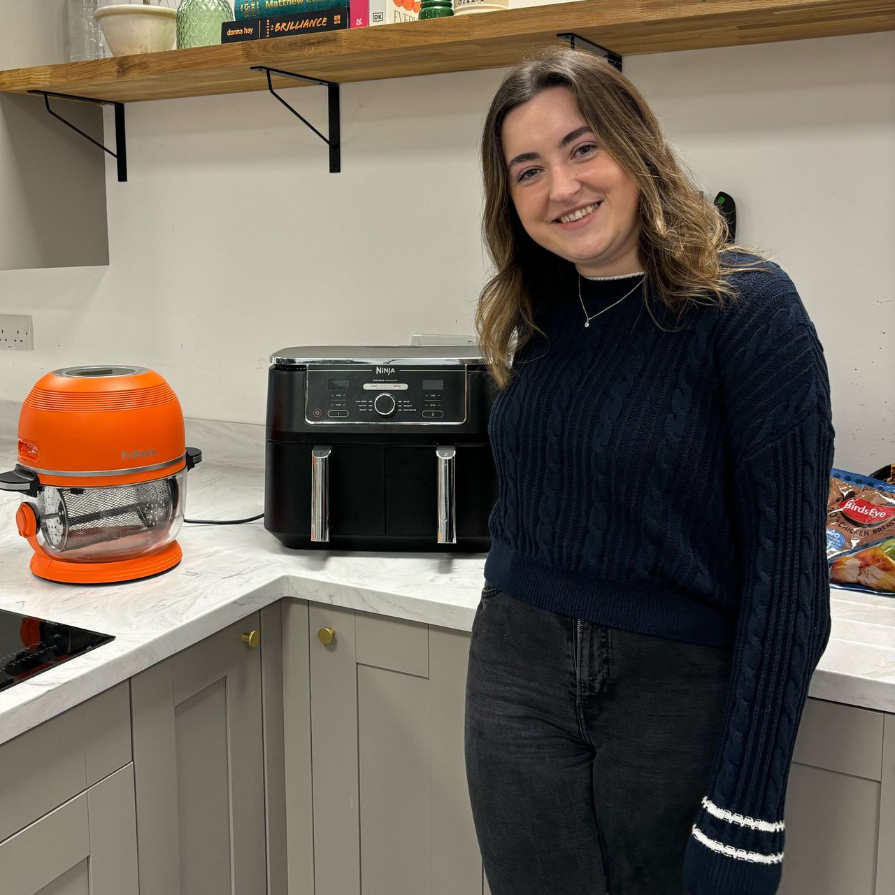 Molly Cleary wearing black jumper and jeans stood in green kitchen testing air fryers on the worktop