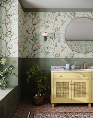 A sage green bathroom with a yellow vanity and bird print wallpaper on the top two thirds of the wall