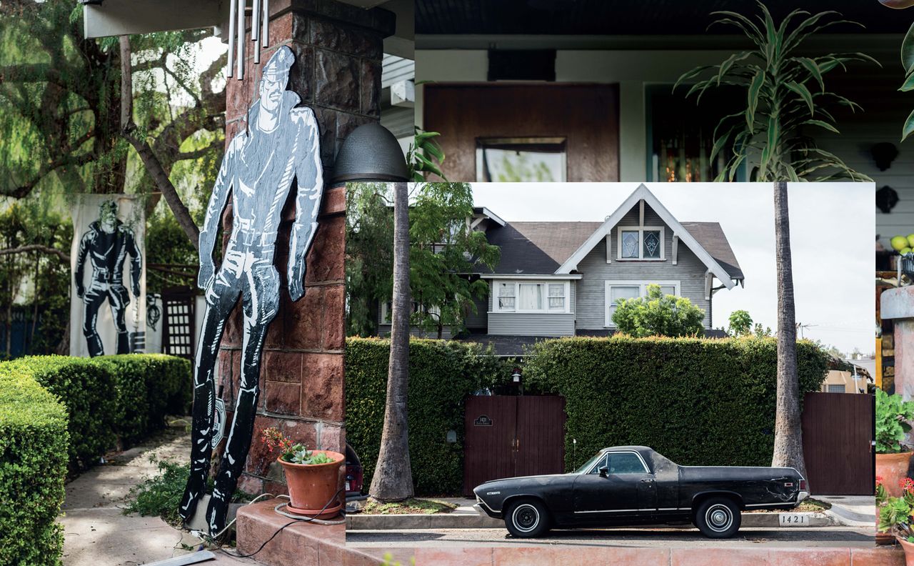 Daytime image outside Tom of Finlands House, Tall hedges, black parked car on road, palm tree trunks, top half of house, tiled roof, windows, backdrop image of the bricked doorway, cardboard cut out black and white male in policeman&#039;s uniform, paved path to the side of the house, trees and small hedges and ceramic plant pot