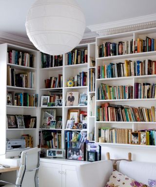 A white home office with a round large white ceiling light, white shelving to the ceiling on two aspects, a white couch, a pine desk and white leather office chair