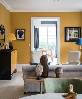 A bright living room painted in yellow with gray sofas, a yellow and black chest of drawers, artwork on the walls, gray floors, and a green chair sat at a marble dining table