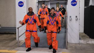 four astronauts with not helmets walk down a ramp from a pair of double doors.