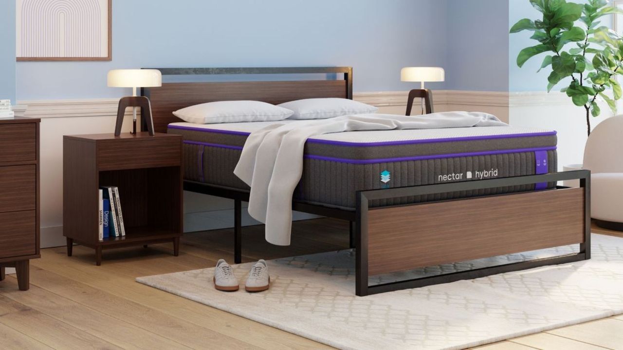 Nectar Premier Hybrid mattress on a wood bed base on a white rug; a lamp and a nightstand either side; a houseplant in the right corner; all against light blue walls. 