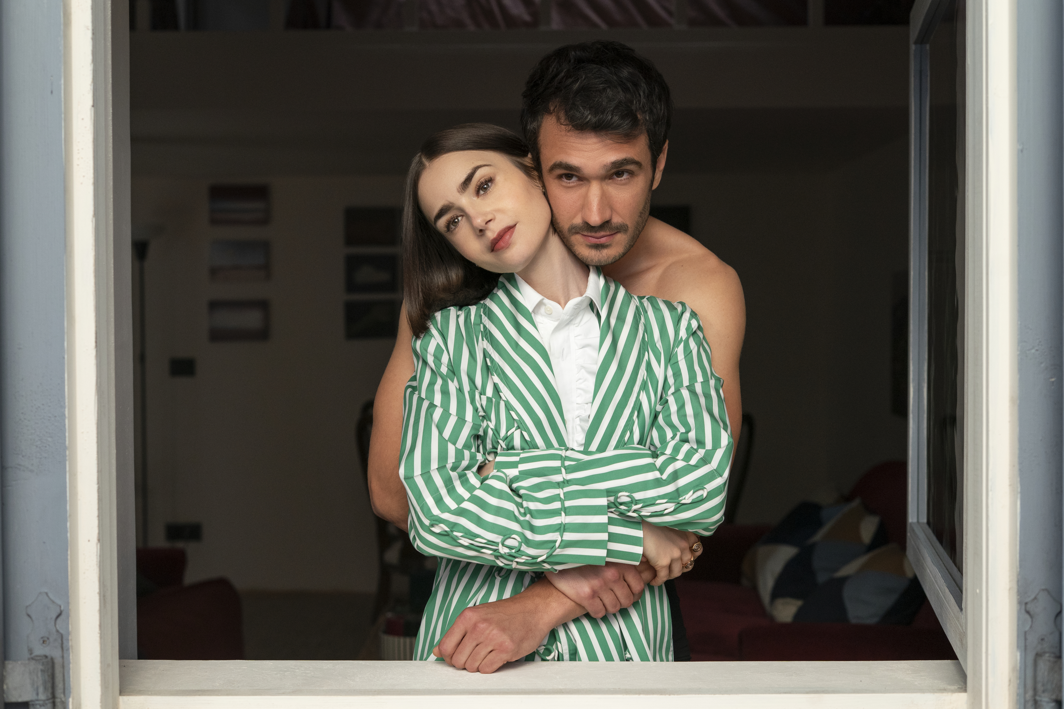 A man (Eugenio Franceschini as Marcello) hugs a woman (Lily Collins as Emily) from behind as they stand in front of an open window, in 'Emily in Paris' season 5.