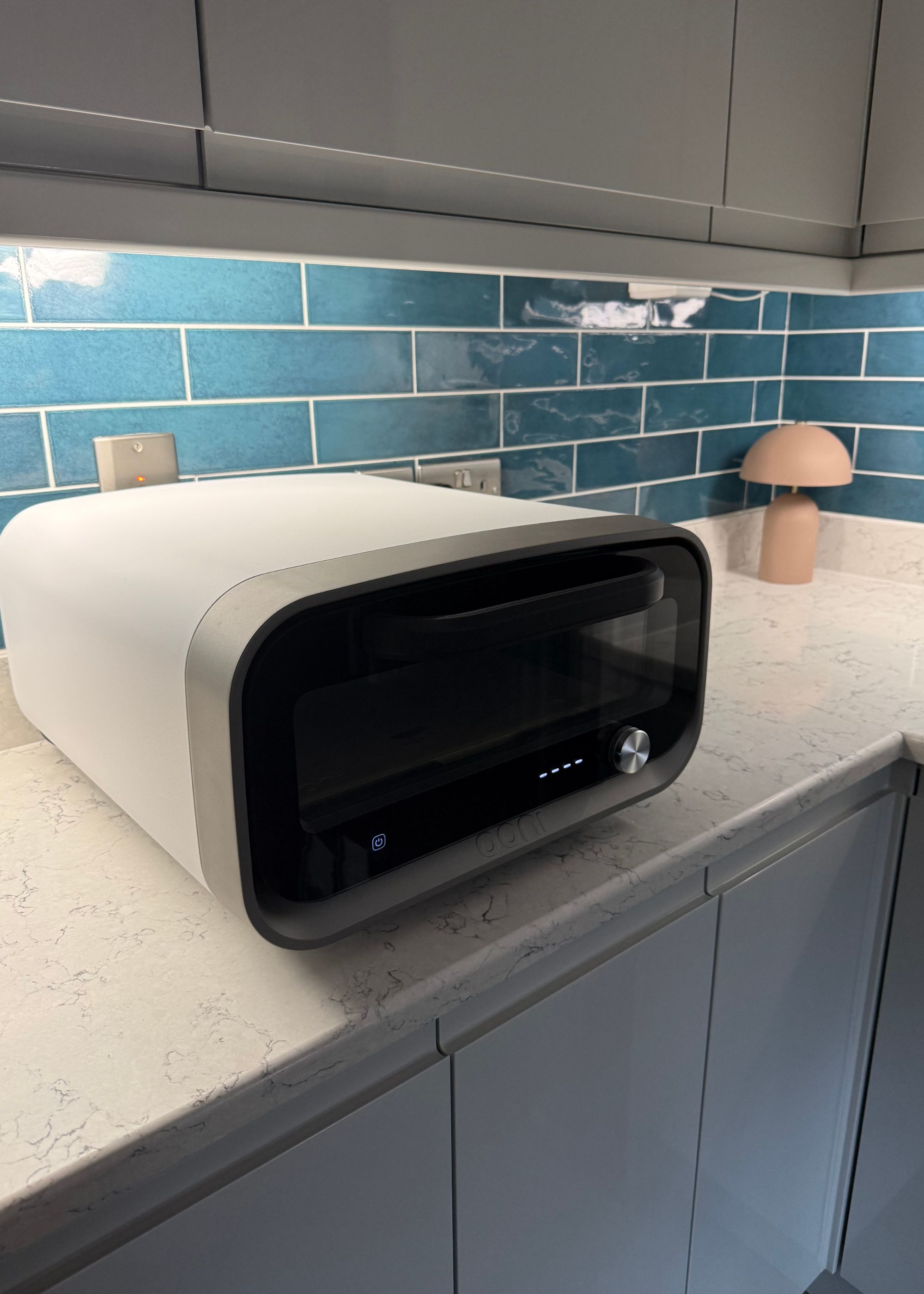 Ooni's volt 2 pizza oven in polar white on a white veined kitchen countertop with a tiled glossy teal backsplash behind it and glossy light gray cabinets above and below it