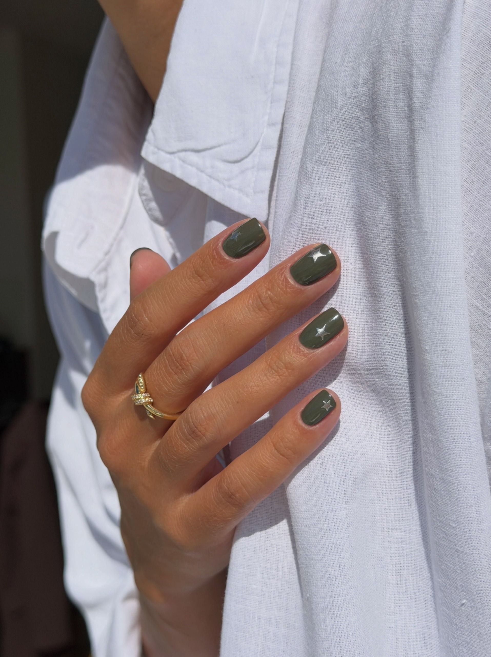 A hand with short olive green nails and tiny silver star accents rests against a white linen shirt