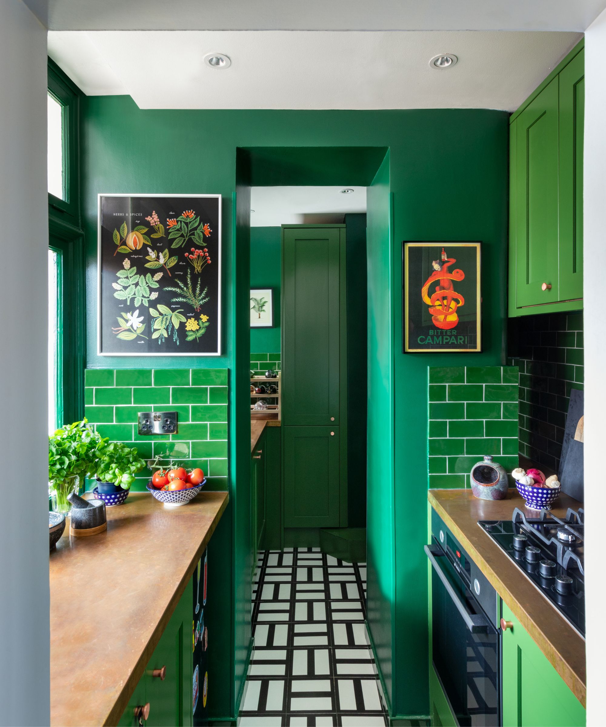Green galley kitchen with bright green painted cabinetry with gold handles, green tiled walls, forest green walls, black and white tiled floor, brushed metal countertops, and Polish pottery