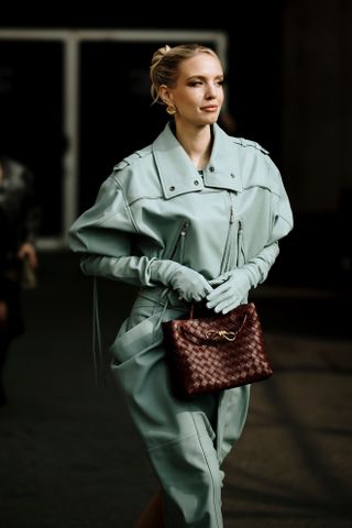 A Milan fashion week guest wearing a blue leather jacket, gloves, and pants carrying a brown Bottega Veneta Andiamo bag