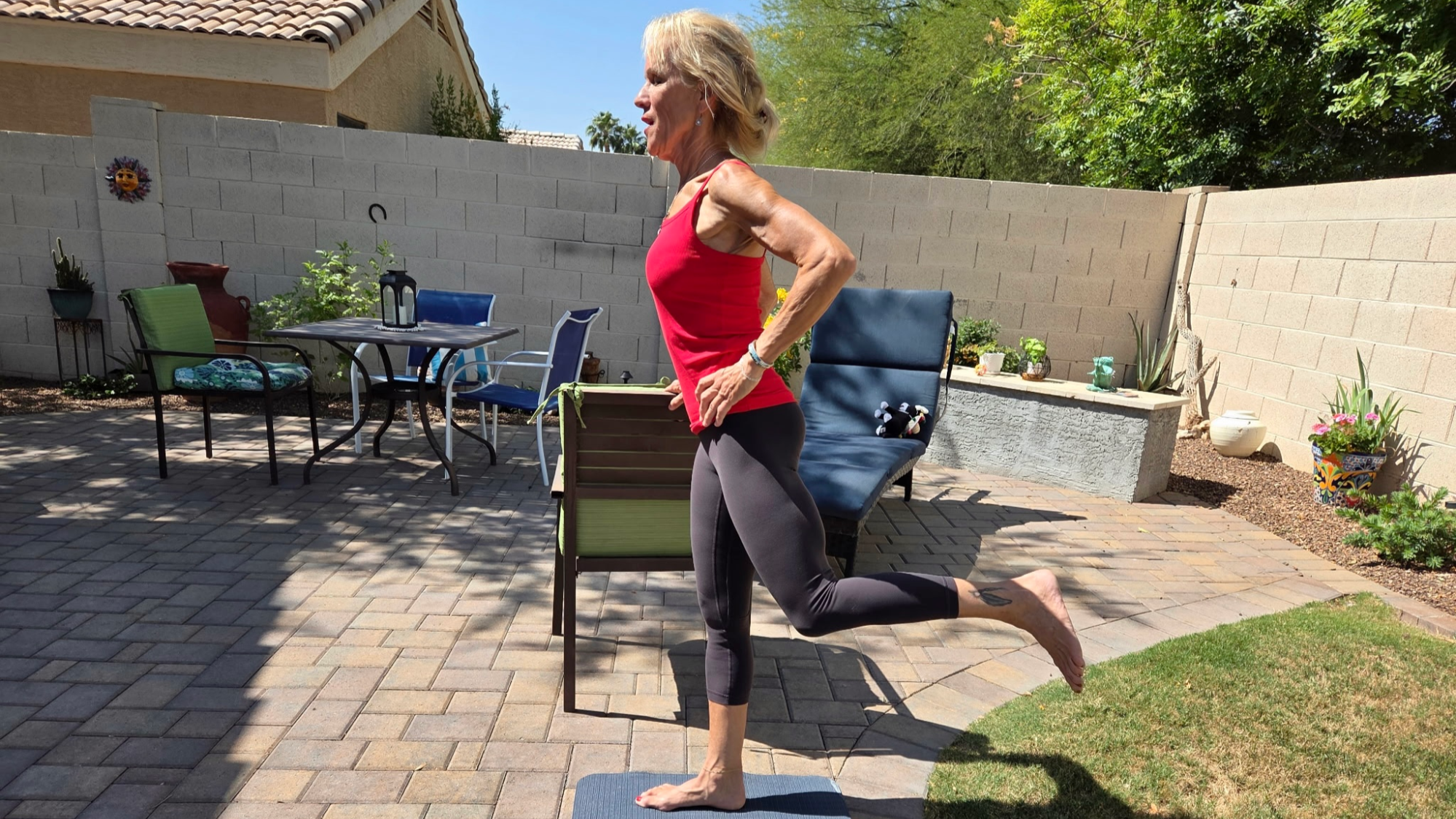 Woman in black leggings and red tank top stands on exercise mat on patio outside, next to grass. She stands side on to a chair, holding it in one hand. One leg is raised behind her.