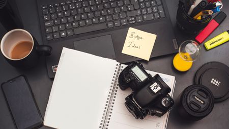 A workspace featuring a laptop, coffee cup, camera, notepad with "Budget Ideas," and colorful stationery on a dark surface