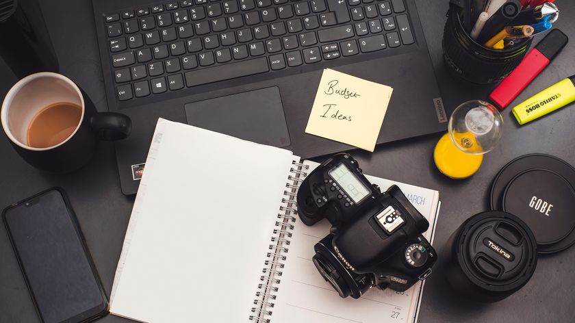 A workspace featuring a laptop, coffee cup, camera, notepad with "Budget Ideas," and colorful stationery on a dark surface