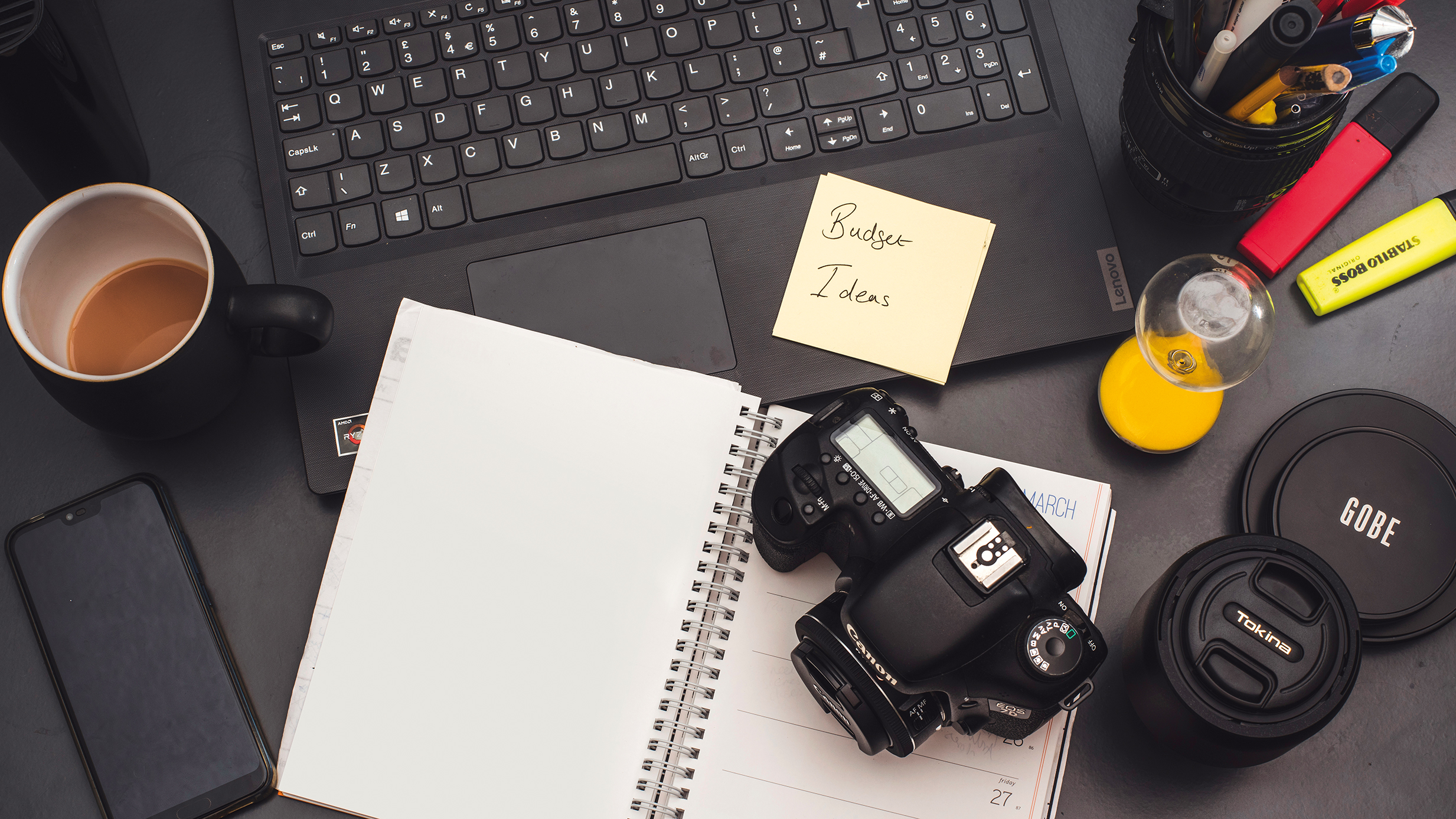 A workspace featuring a laptop, coffee cup, camera, notepad with "Budget Ideas," and colorful stationery on a dark surface