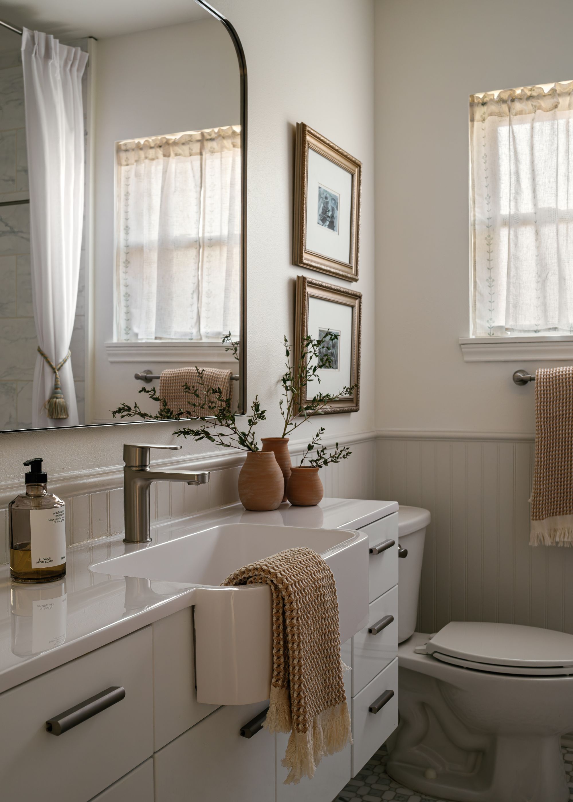 A bathroom with a quartz countertop and a large sink with a towel hanging from it. Above the sink basin is a large mirror. There is also a view of the toilet with art frames hanging above it.