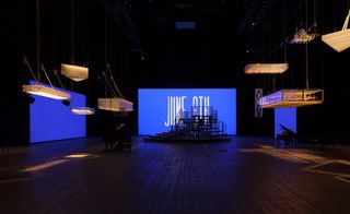 Large metal light structures hanging in an exhibition hall with a grand piano and blue multi-media screens