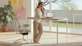 A woman with a laptop standing at the Desktronic HomeMax desk with a SitPro office chair behind her