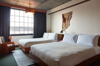 Ace Hotel Brooklyn room, Tamika Rivera textile art on the wall over two double beds, cream covers, white sheets and pillow, dark grey curtains, black frame window, wooden ceiling lamp, striped wooden rocking chair, grey marble ceiling, white walls, grey rug under bed