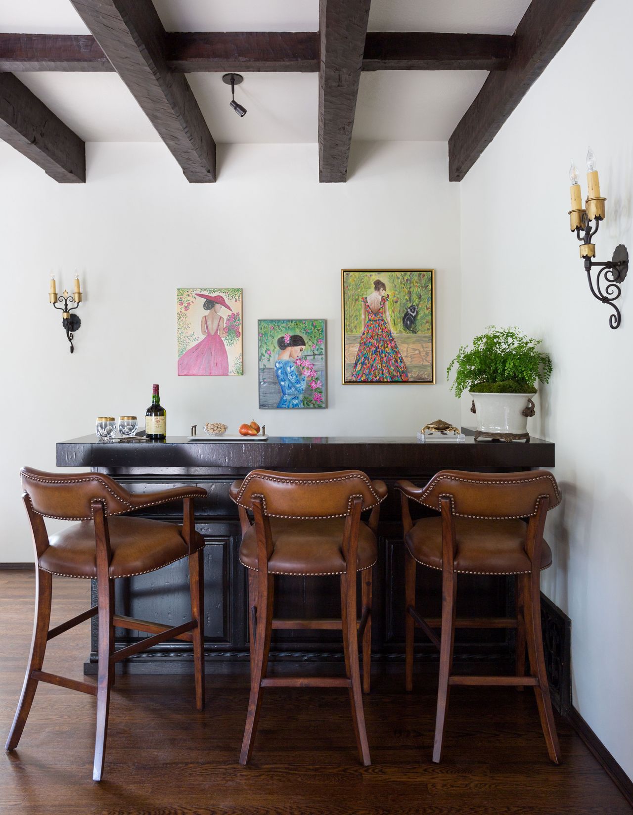 custom bar with ceiling beams above and three leather bar stools and bright pictures behind and dark wood counter with drinks bottle on and leaded windows behind and dark ceiling beams
