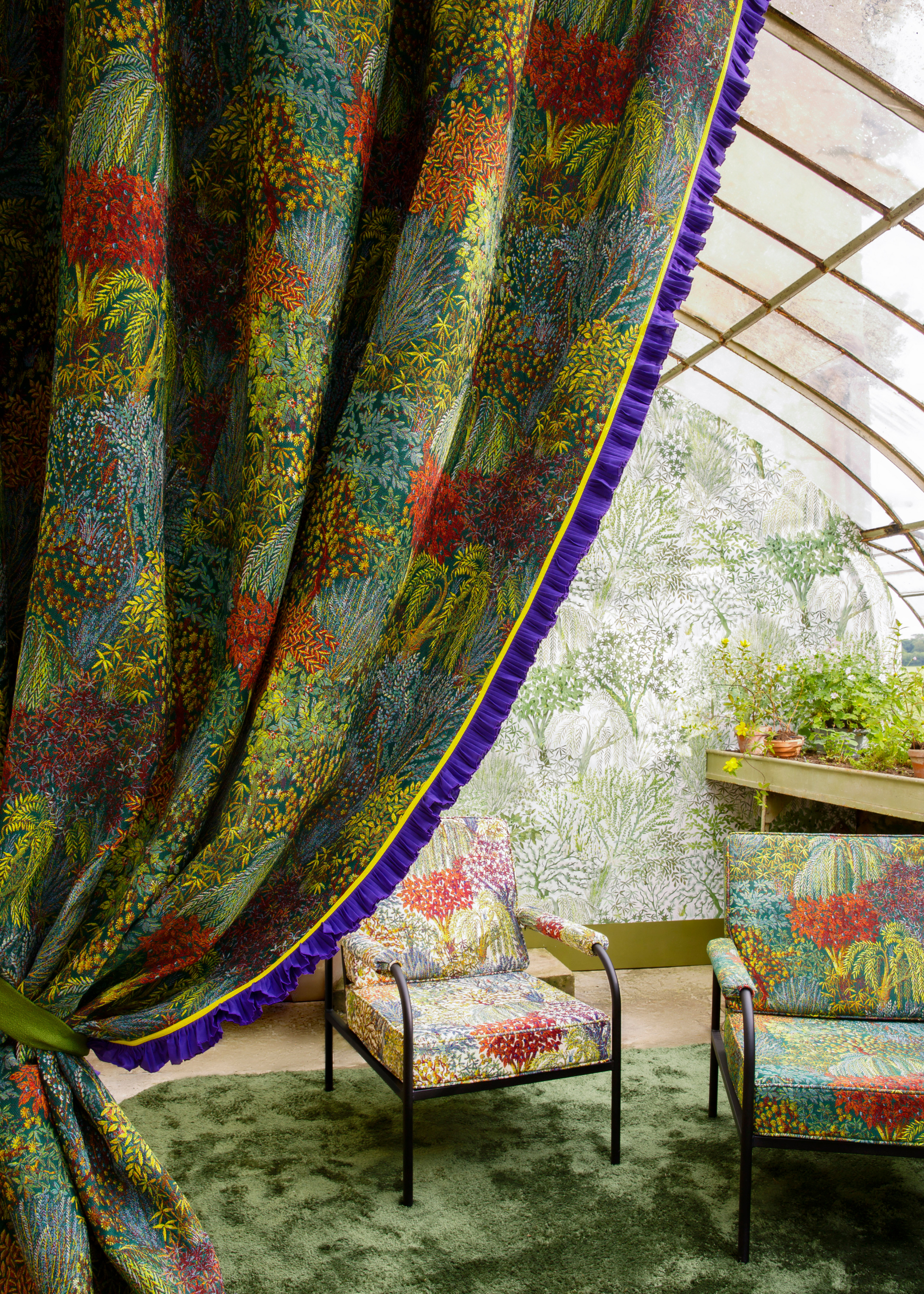 A close-up of a floral curtain tucked away leading to a greenhouse with a verdant wallpaper, a green area rug and a pair of chairs with floral upholstery