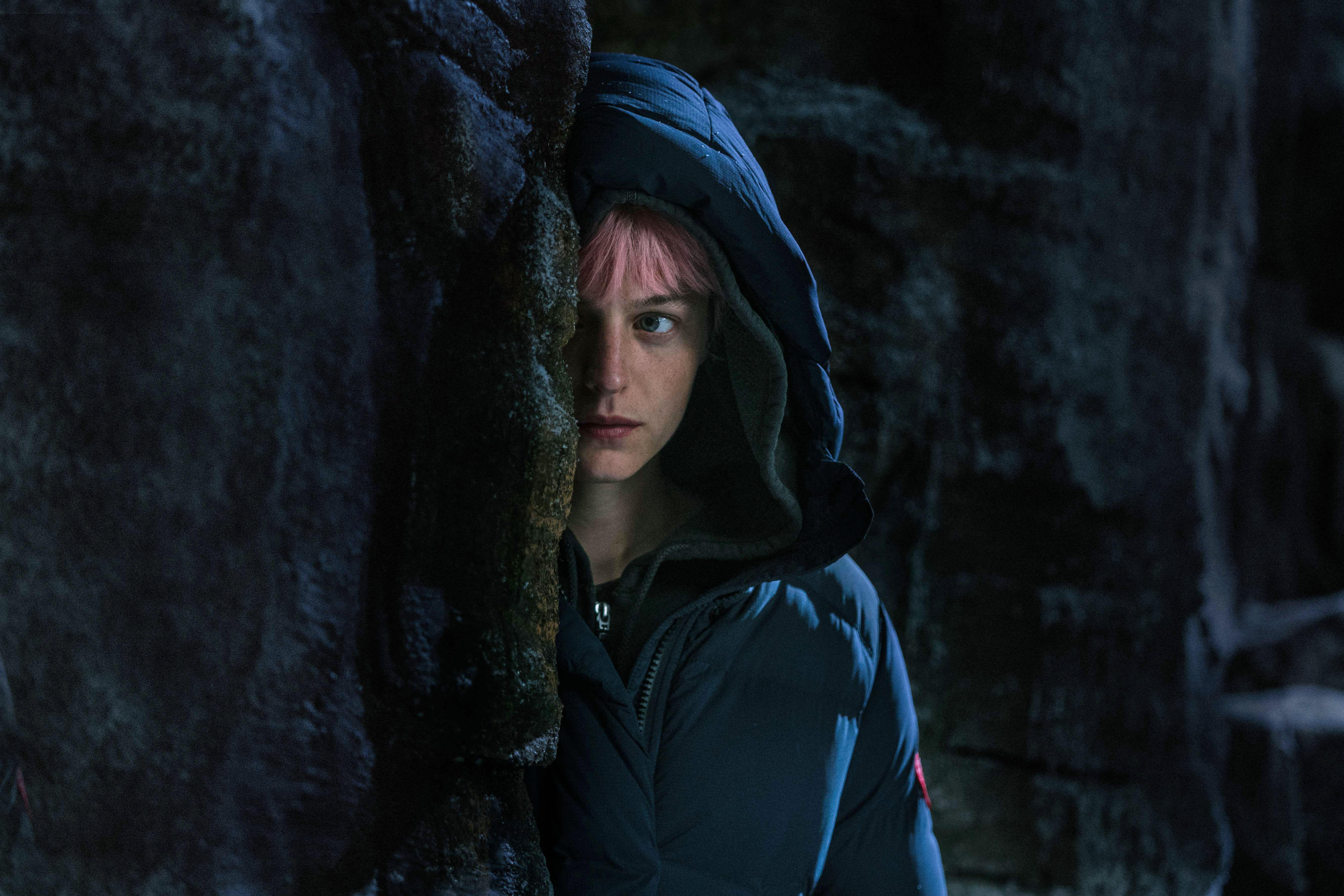 emma corrin wears a sweatshirt and peers around a corner in a still from murder at the end of the world