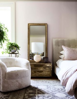 a bedroom with plants, an upholstered bedhead, light sheets, a fluffy armchair and an antique gold bedside table with a mirror on top