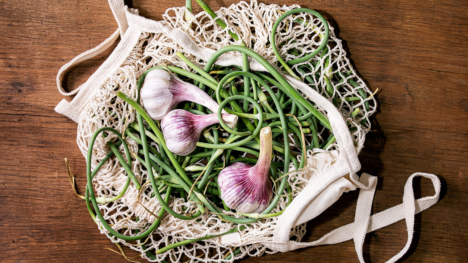 Garlic bulbs and garlic scapes in mesh grocery bag on wooden counter