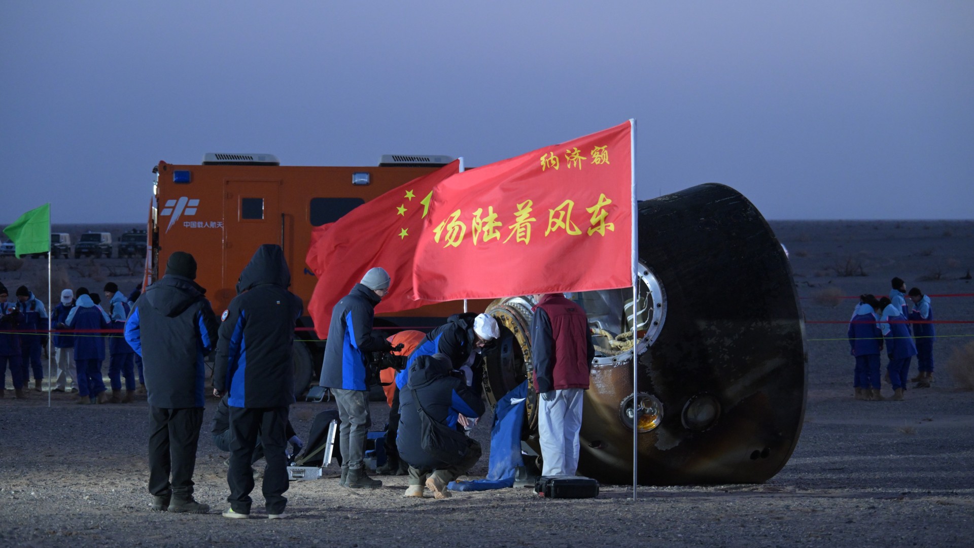 a scorched cone-shaped capsule lies on a desert floor surrounded by people in thick blue coats