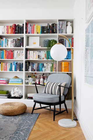 A corner of the living room with black and grey armchair, gold and marble floor lamp, and a wall of bookshelves decorated with bottles