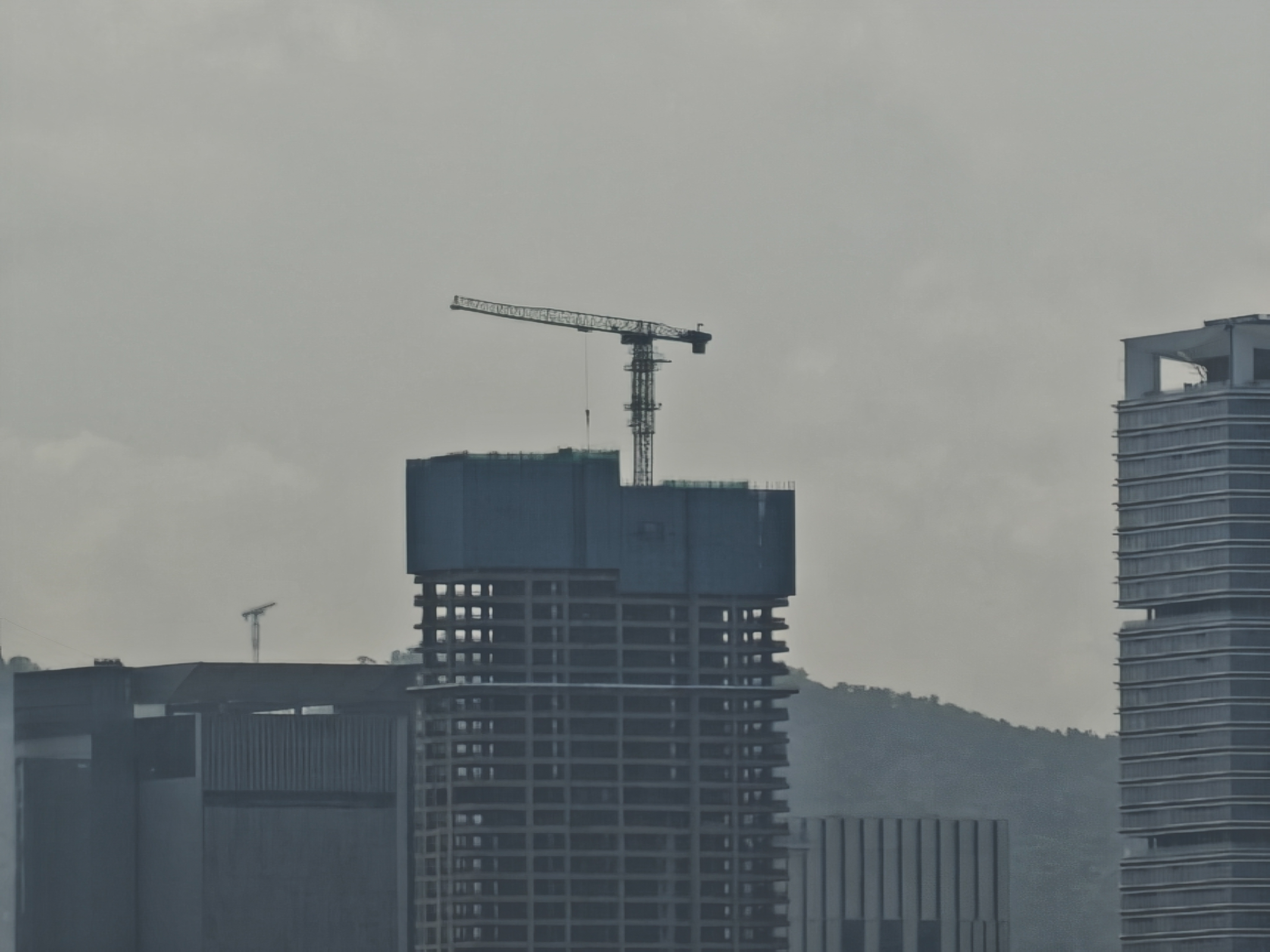 A digitally zoomed-in shot of a skyscraper under construction. A large construction crane is perched on top of the dark, unfinished concrete structure. The image has a slightly grainy, painterly texture due to the high zoom level, and the background shows the silhouette of a mountain under a gray sky.