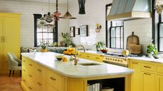 yellow kitchen cabinets with white marble counter, different pendant lights hanging overhead, a freestanding oven with chopping boards leaning against the white tile backsplash