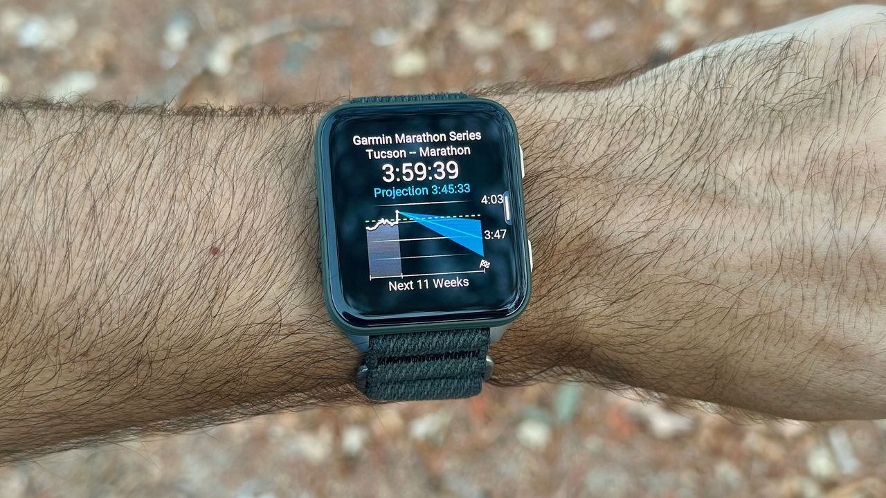 The Garmin Venu X1 on the author's wrist showing the Primary Race Predictor widget, with a current and projected marathon race time for the Garmin Marathon Series in Tucson in 11 weeks.