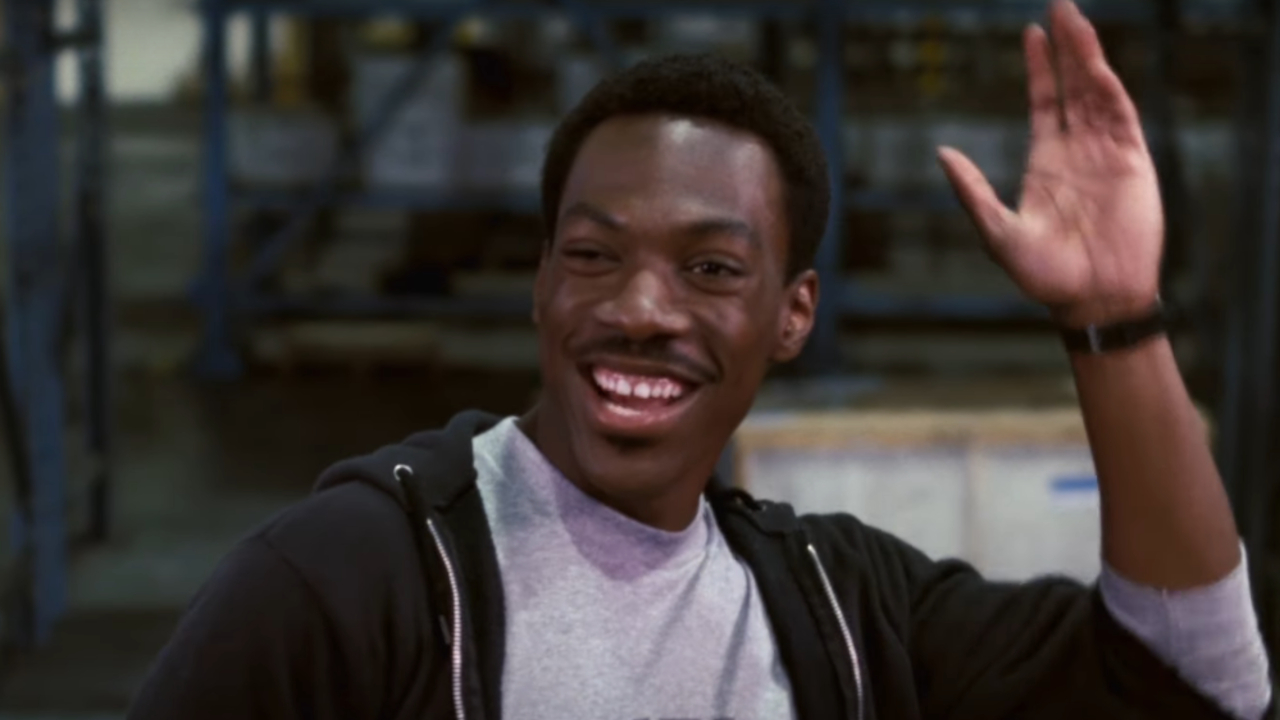Eddie Murphy waves while smiling in a warehouse in Beverly Hills Cop.