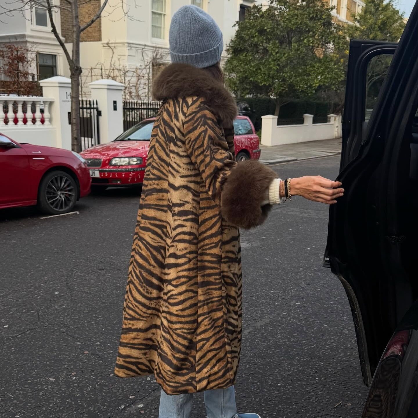 Woman wears a fur lined leopard print coat, blue beanie, light wash jeans and blue Vans slip ons. 