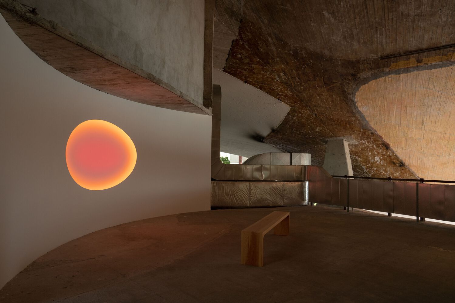 A softly glowing, organically shaped sculptural installation resembling the sun sits on an arched, bare white wall inside a modernist building punctuated by rock, wood, and cement surfaces.