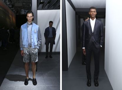 Two side-by-side photos of male models wearing looks from Brioni's collection. In the first photo there is a model wearing a grey top, blue and grey jacket, grey patterned shorts and black shoes. In the background there is a man wearing a shirt, trousers and coat. In the second photo there is a model wearing a white shirt, black suit and black shoes. In the background there is a man facing in a different direction