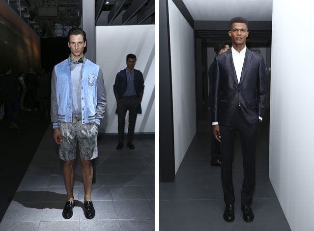 Two side-by-side photos of male models wearing looks from Brioni&#039;s collection. In the first photo there is a model wearing a grey top, blue and grey jacket, grey patterned shorts and black shoes. In the background there is a man wearing a shirt, trousers and coat. In the second photo there is a model wearing a white shirt, black suit and black shoes. In the background there is a man facing in a different direction