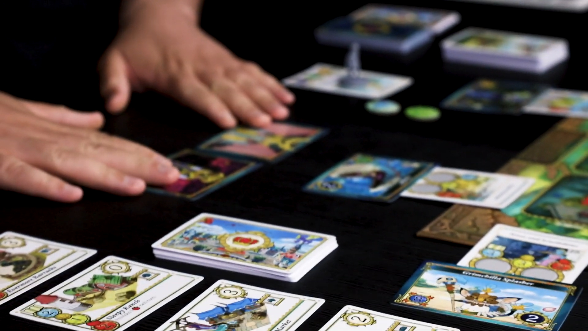 Two hands laid on a dark surface behind cards, tokens, and components from Ni No Kuni 2: The Board Game