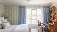 Bedroom with blue and green floral wallpaper, a large window with blue curtains, a mahogany desk filled with books and a chair underneath, and a white wooden bed with bed linen and floral cushions.